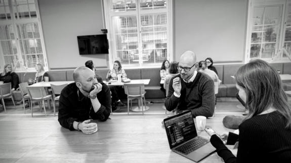 Kafé-miljö med tre personer i fokus, engagerade i diskussion, omgiven av andra som arbetar och pratar.