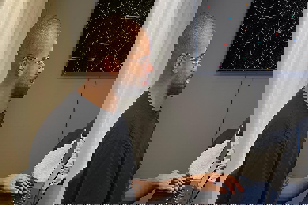 En man sitter vid ett skrivbord och arbetar på en laptop, med en grafisk skärm i bakgrunden.