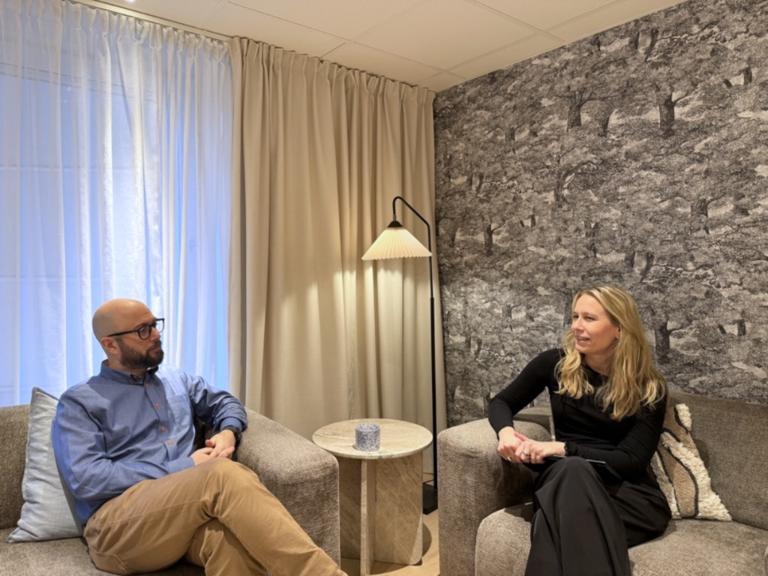 En man och en kvinna diskuterar i en stilren miljö med texturerade väggar och naturligt ljus.