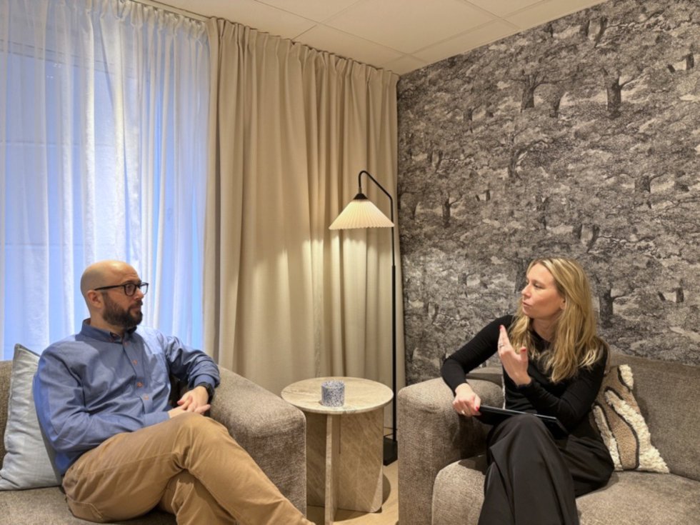 Två personer sitter i en modern, ljus miljö och diskuterar, omgivna av mjuka texturer och en lampa.