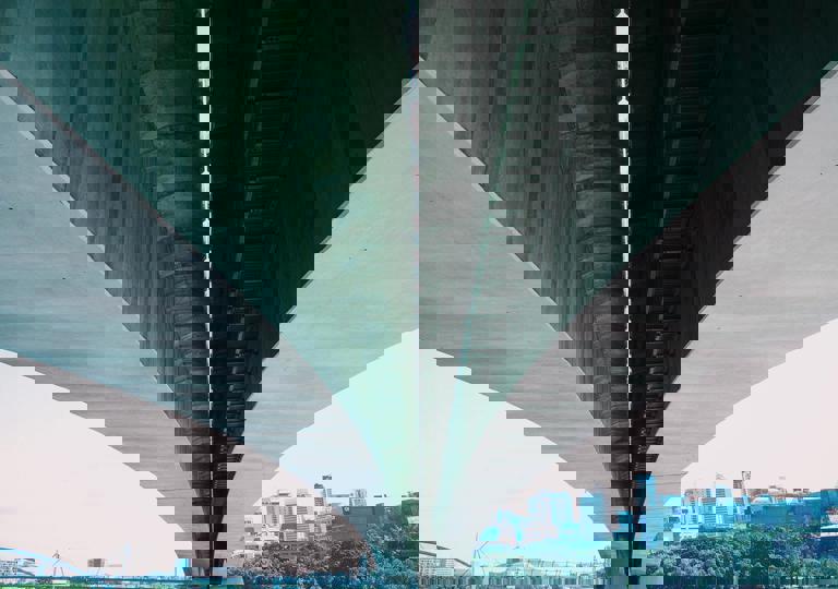 Perspektiv av en bro med två parallella sektioner, omgiven av stadslandskap och grönska.