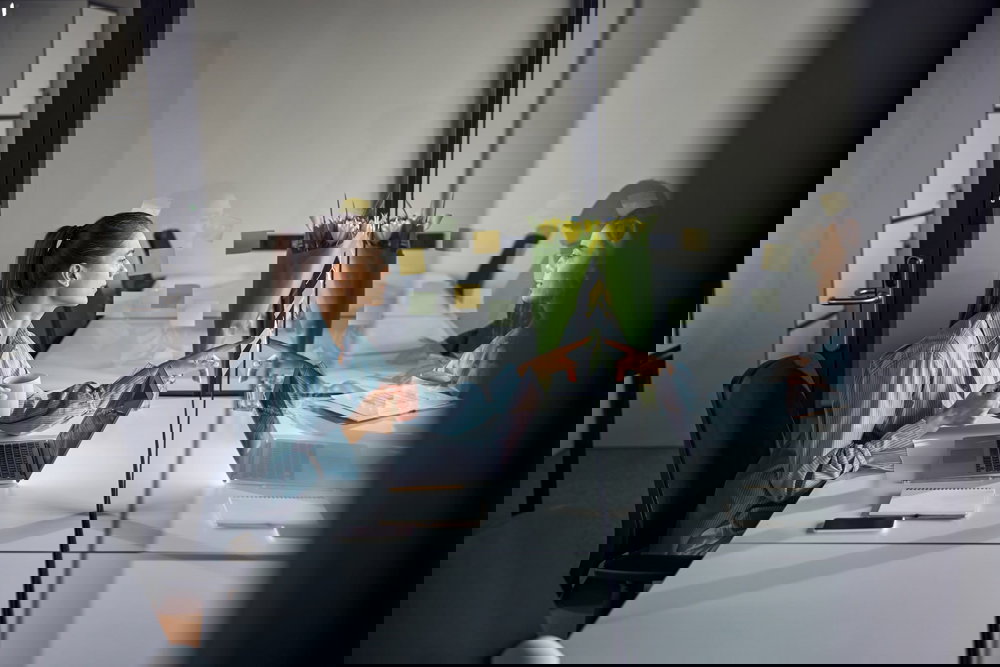 Kvinna med laptop och kopp, granskar sina anteckningar i ett modernt kontorsutrymme.