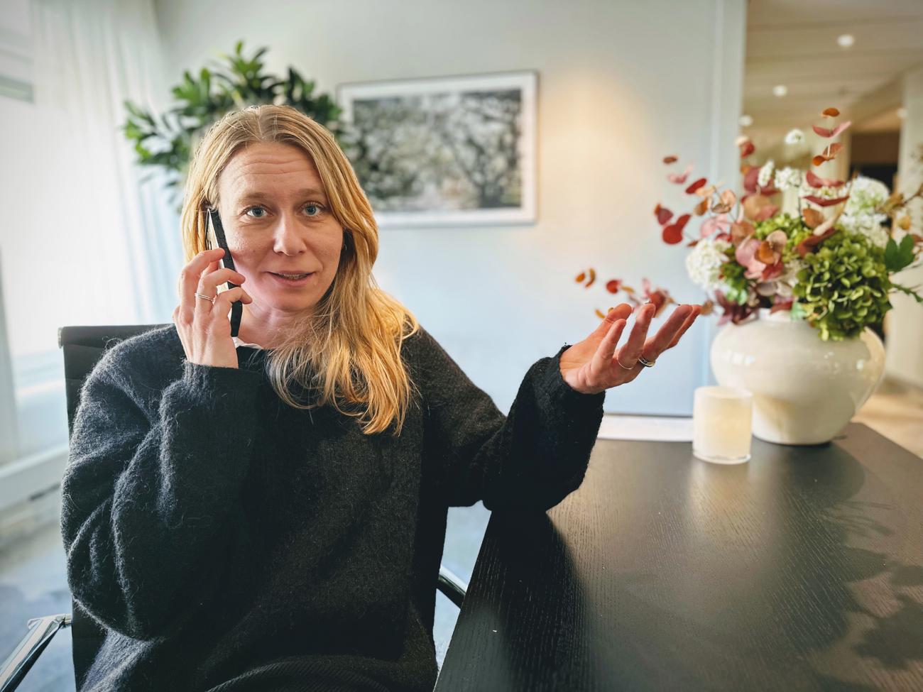 Kvinna gestikulerar medan hon pratar i telefon, sittande vid ett skrivbord med en ljus blomsterarrangemang.