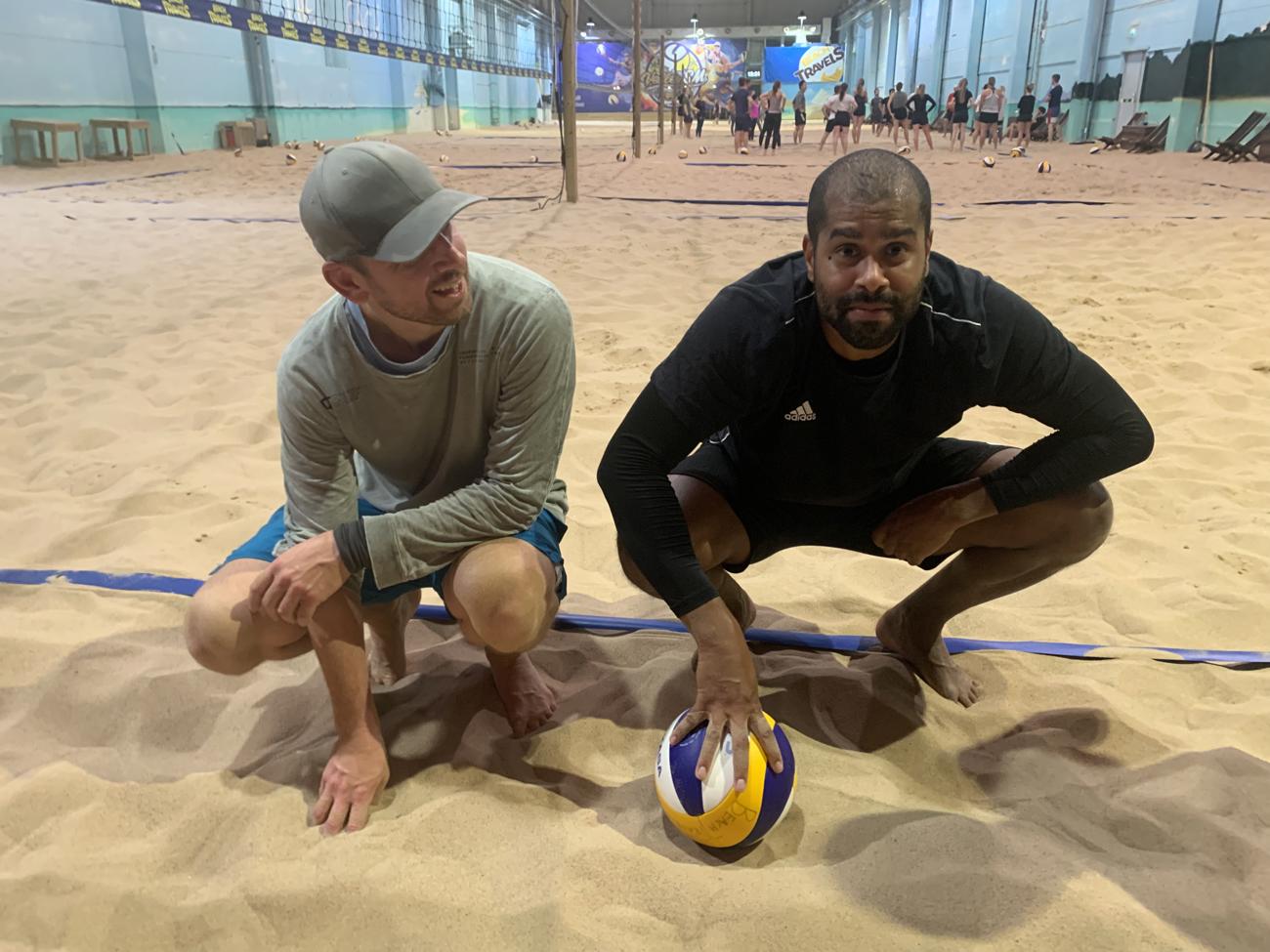 Inomhus volleybollspelare i sand, en med volleyboll, den andra ler mot kameran.