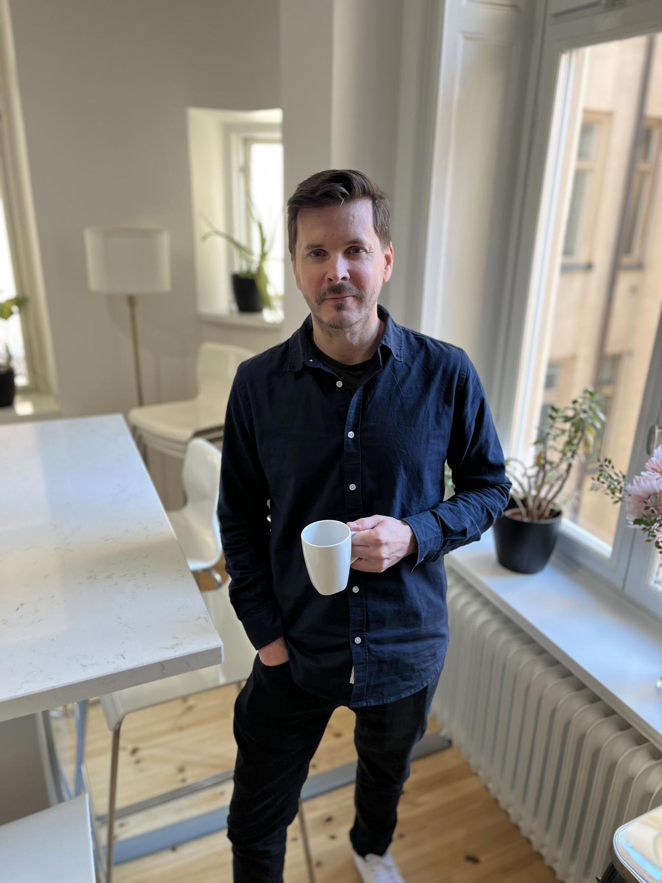 Man står vid ett fönster i ett ljust kontor, håller en vit kopp och ser mot kameran.