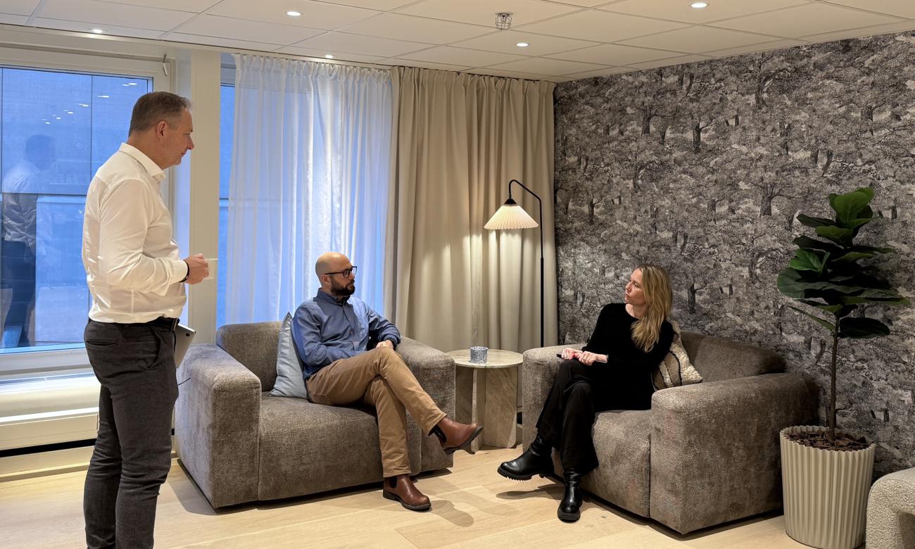En man står och pratar med två sittande personer i en inbjudande kontorsmiljö med mjuka möbler och naturligt ljus.